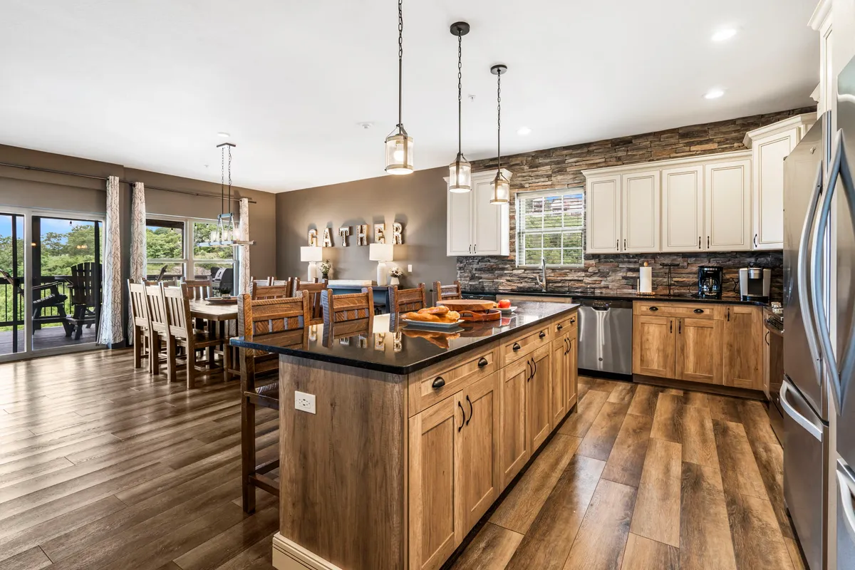 An open-concept kitchen and dining area with a central island, a large dining table, stone accent wall, and 'GATHER' lettering. The space is filled with natural light and arranged for both meal preparation and group dining.