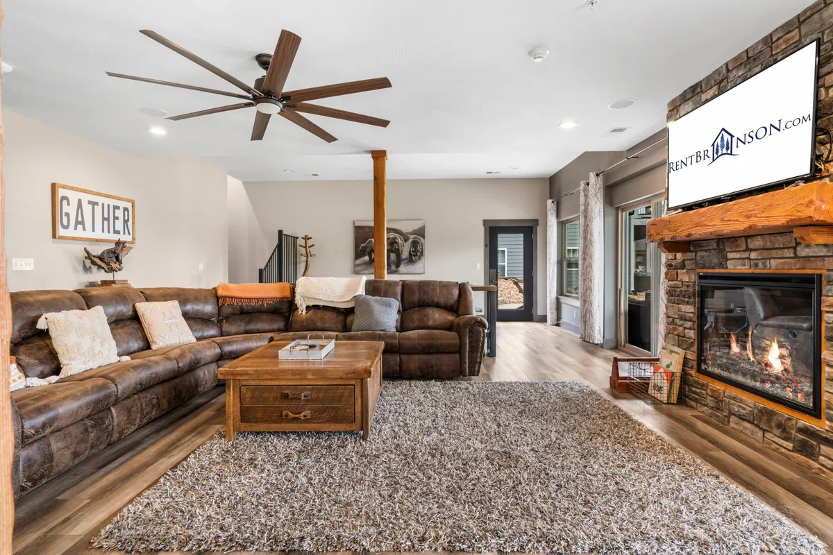 A spacious and inviting living area featuring a large sectional sofa, a cozy fireplace, coffee table, television, and prominent 'GATHER' sign. The room is designed for group activities like movie nights and socializing, with ample seating and warm lighting creating a communal atmosphere.