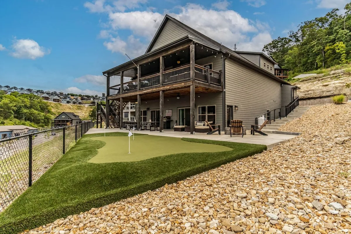 This bright exterior shot showcases the deck area overlooking the private 4-hole putting green, with the house facade and outdoor lounging spaces visible. The green is set against a backdrop of lush landscaping, ideal for friendly family competitions and group gatherings.