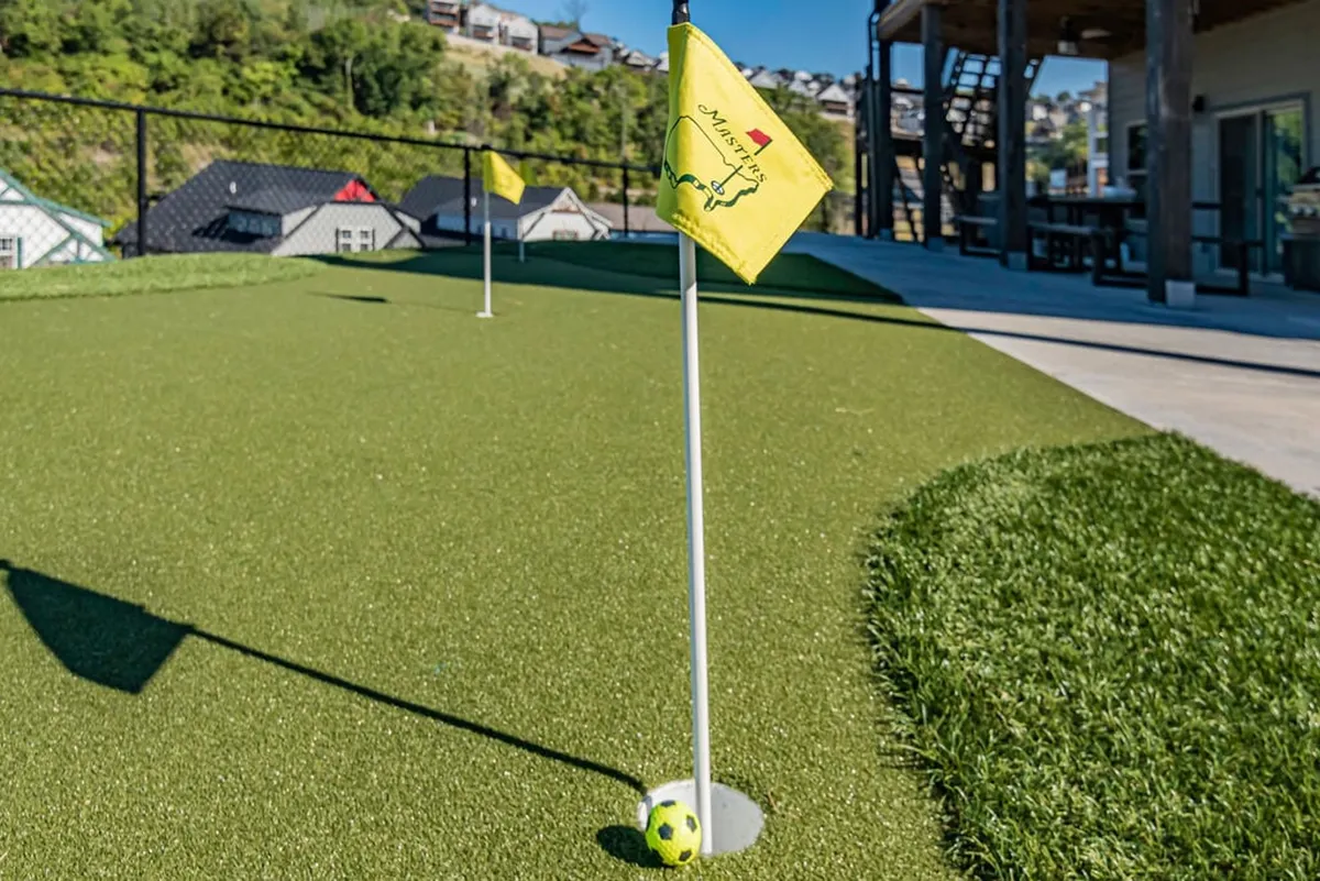 A vibrant outdoor putting green with golf flags and a soccer ball set on neatly manicured grass, surrounded by open sky and a hint of gathering space for friendly games.