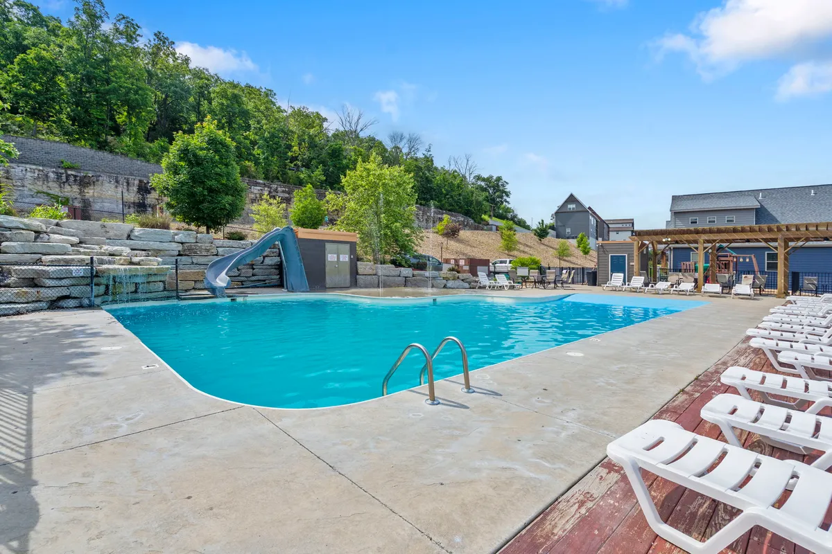 The vibrant outdoor pool scene showcases a sparkling pool surrounded by lounge chairs, a water slide, and lush landscaping. Families and friends gather poolside, with children enjoying playful moments and adults relaxing in a joyful, sunny setting.