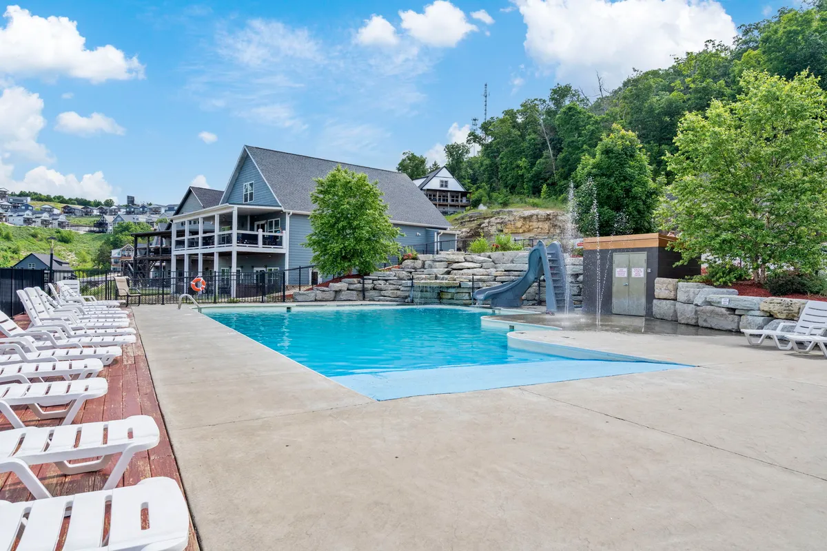 A lively outdoor resort pool featuring a water slide, lounge chairs, and landscaped trees, with houses in the background providing a community ambiance.