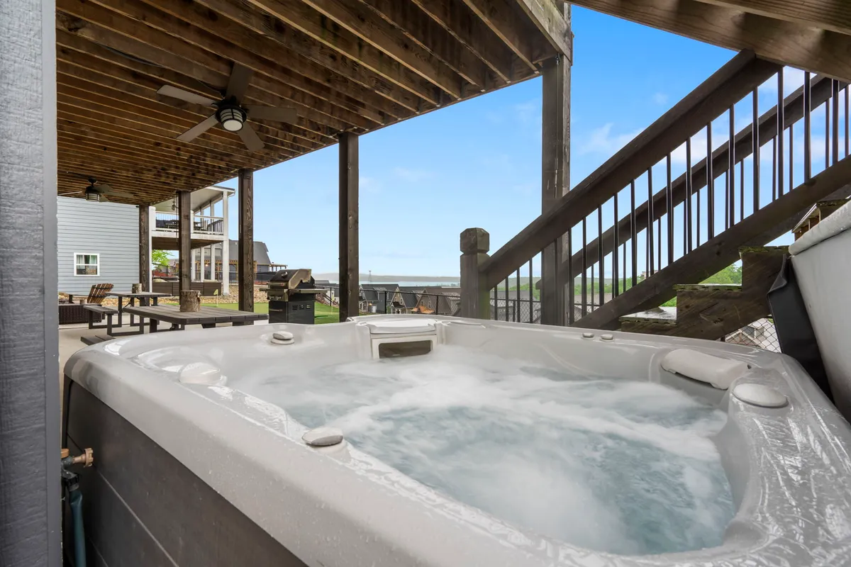 The image focuses on the inviting hot tub nestled under a wooden structure, a staircase leading to a spacious outdoor seating area, and a serene natural backdrop. It suggests relaxation and unwinding in comfort, regardless of the weather.